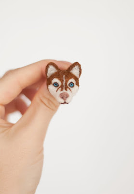 A wool pin in the shape of the head of a red and white Siberian husky with blue eyes